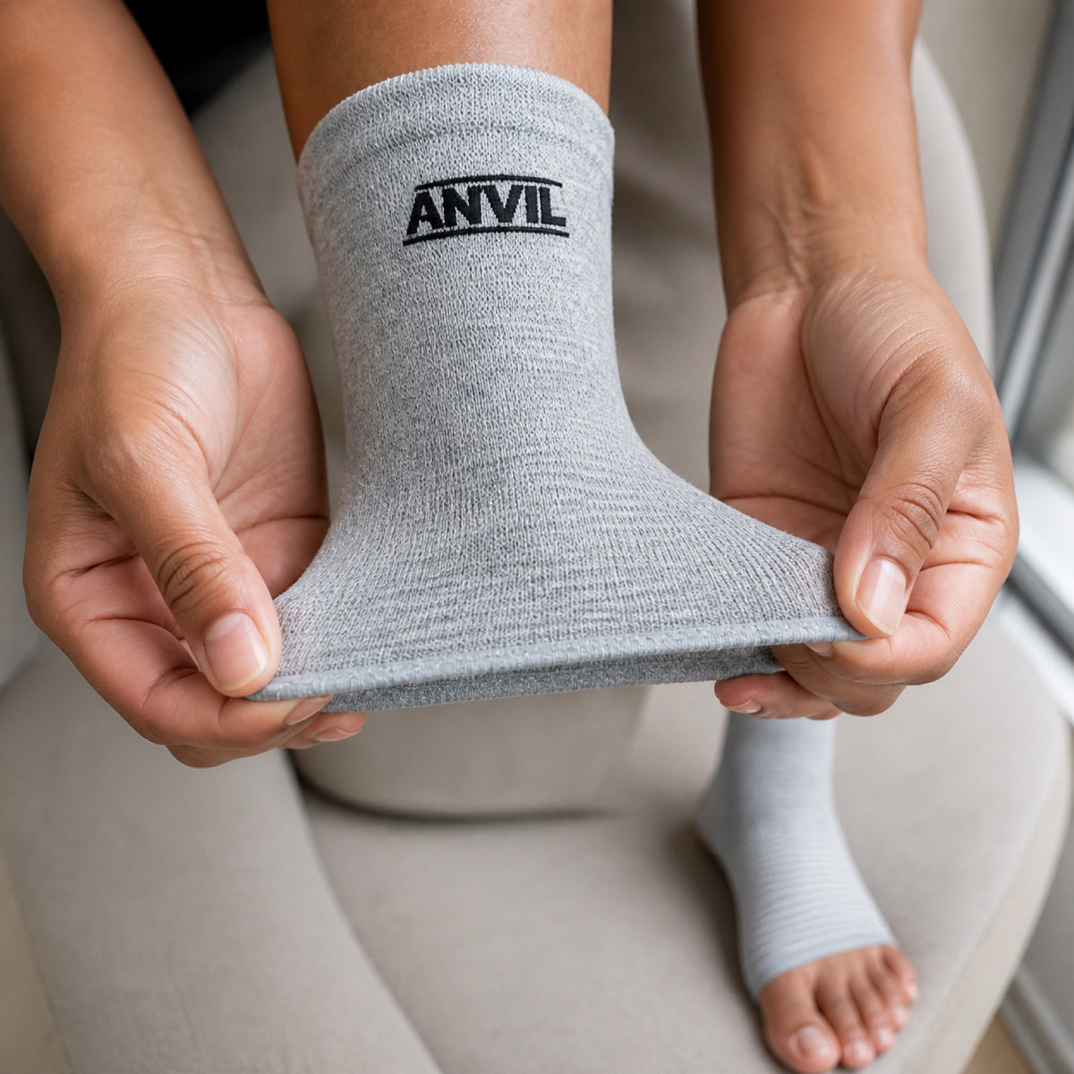 Two hands stretching the open toe edge of a gray ANVIL foot sleeve, the other foot already wearing one.