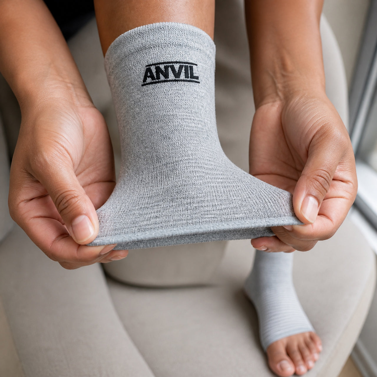 Two hands stretching the open toe edge of a gray ANVIL foot sleeve, the other foot already wearing one.