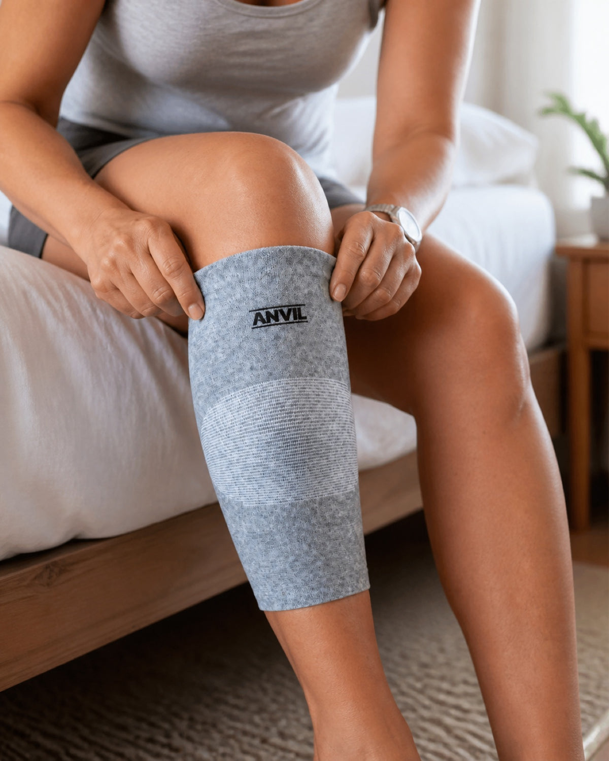 Woman sitting on the edge of a bed pulling a gray ANVIL sleeve up over her calf.