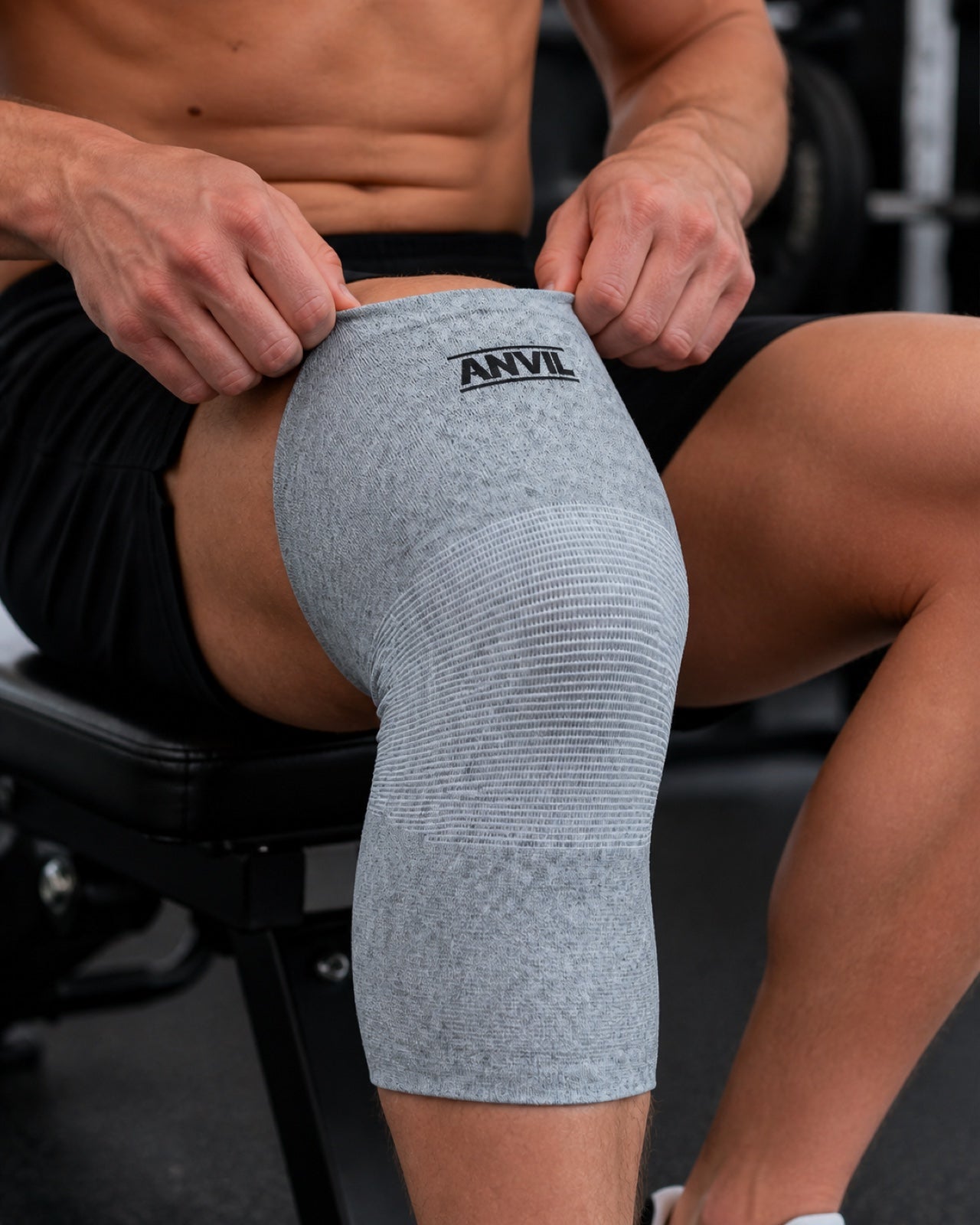 Man seated on a gym bench pulling a gray ANVIL knee sleeve up over his knee with both hands.