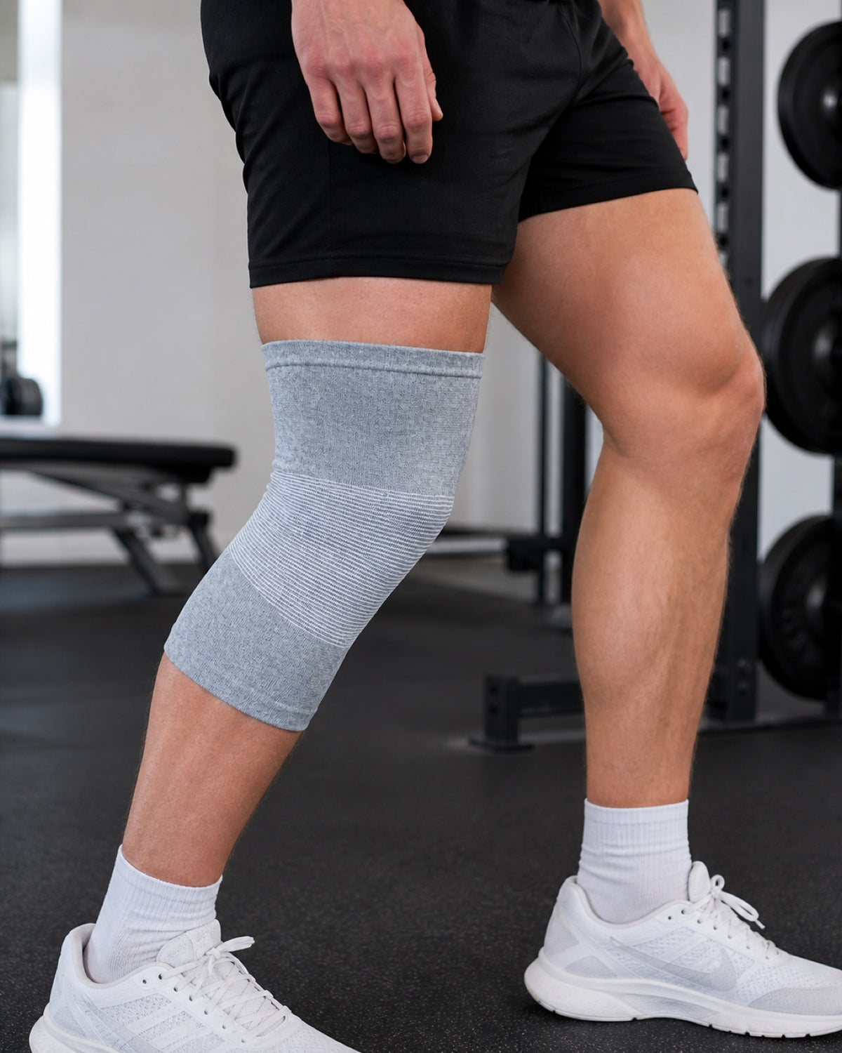 Close view of a man standing in a gym beside a weight rack, with a gray knee sleeve on his left knee.