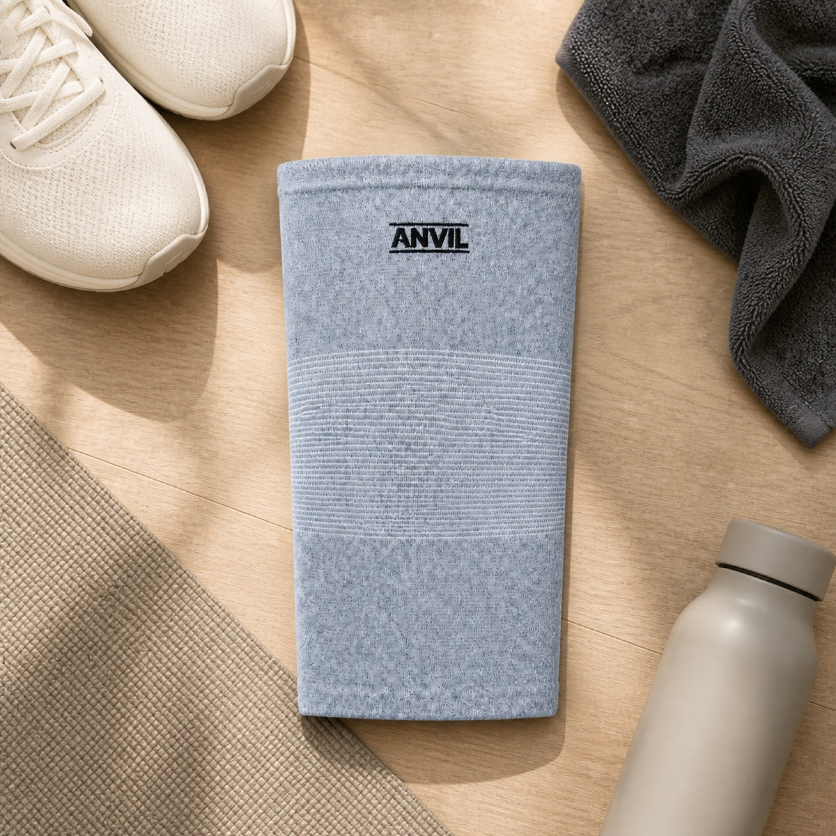 Gray knitted ANVIL knee sleeve laid on a wood floor beside sneakers, a folded towel, a yoga mat and a water bottle.