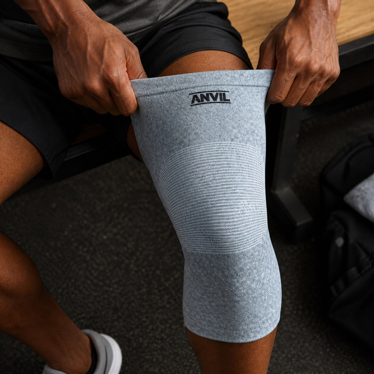 Close view of a man seated on a gym bench pulling a gray ANVIL knee sleeve up over his knee with both hands.
