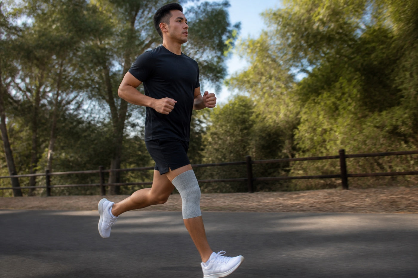 Man running on a paved path lined with trees, with a gray knee sleeve on his left knee.