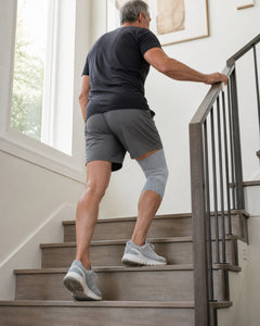 Man climbing a staircase at home, seen from behind, with a gray knee sleeve on his right knee.